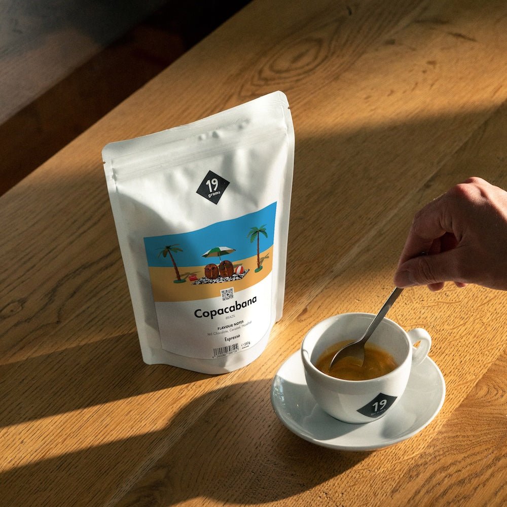 19grams Kaffeeverpackung „Copacabana" Espresso mit tropischer Strandillustration auf Holztisch neben einer Espressotasse