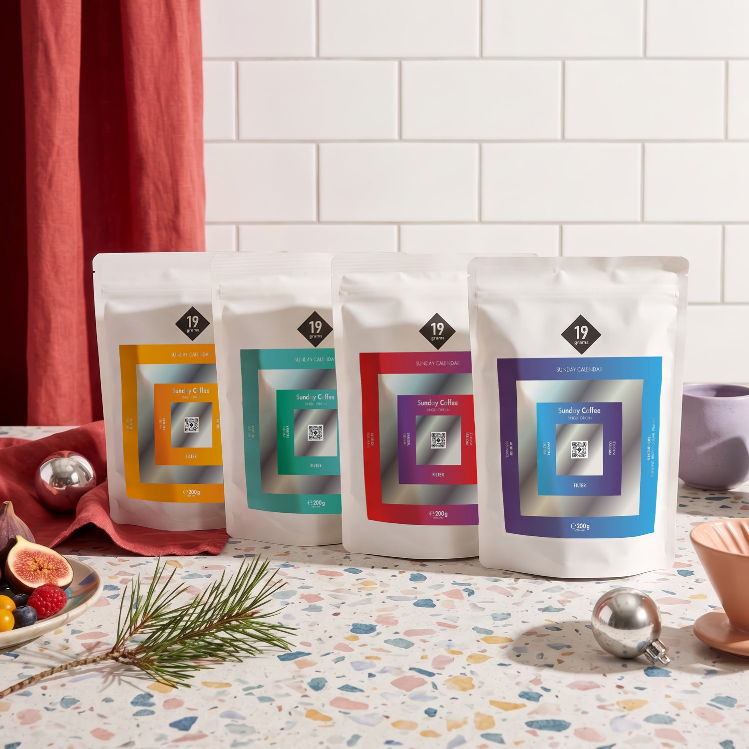 19grams Sunday Calendar Specialty Coffee Moodbild – Vier bunte 19grams Sunday Coffee Kaffeebeutel auf Terrazzo mit Feigen, Beeren und Tannenzweig, Kaffeerösterei Berlin