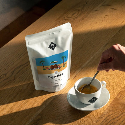 19grams Kaffeeverpackung „Copacabana" Espresso mit tropischer Strandillustration auf Holztisch neben einer Espressotasse