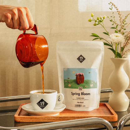 19grams Spring Bloom Specialty Coffee Filter – Kaffee wird aus Karaffe in Tasse gegossen neben Kaffeebeutel und Blumenstrauß, Kaffeerösterei Berlin