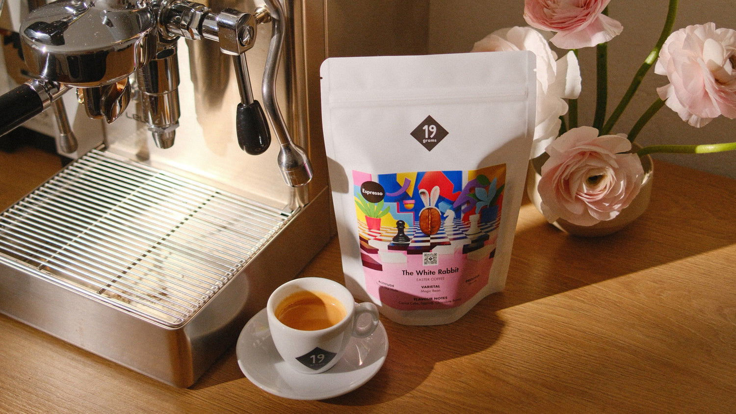 19grams Osterkaffee „The White Rabbit" neben Espressomaschine – Banner mit rosafarbenen Ranunkeln