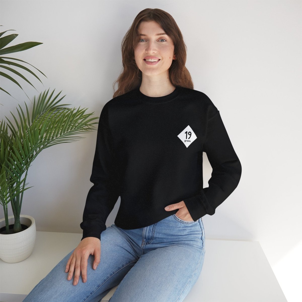 19grams schwarzer Pullover mit weißem 19grams Logo auf der linken Brust, getragen von einer Frau, die auf einem Tisch sitzt