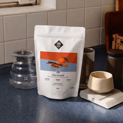 19grams Finca Naranjal Filter Kaffeebeutel neben Glaskaraffe und Keramiktasse auf blauem Tresen – Specialty Coffee Peru Moodshot
