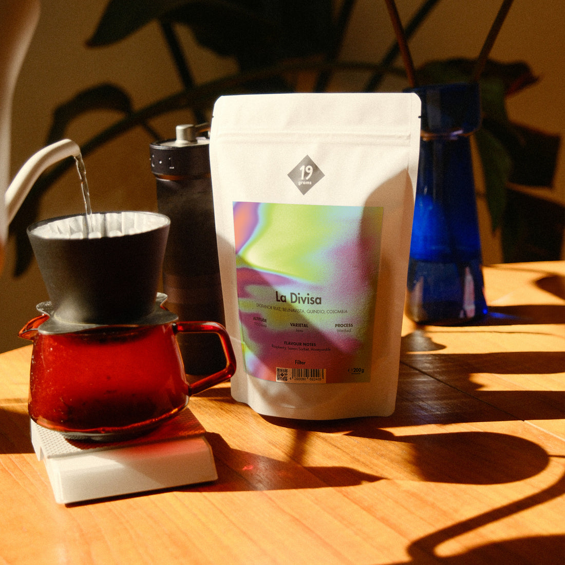19grams La Divisa Specialty Coffee Moodbild – Kaffeebeutel mit Handfilter, roter Tasse und Kaffeezubehör auf Holztisch im Sonnenlicht, Kaffeerösterei Berlin