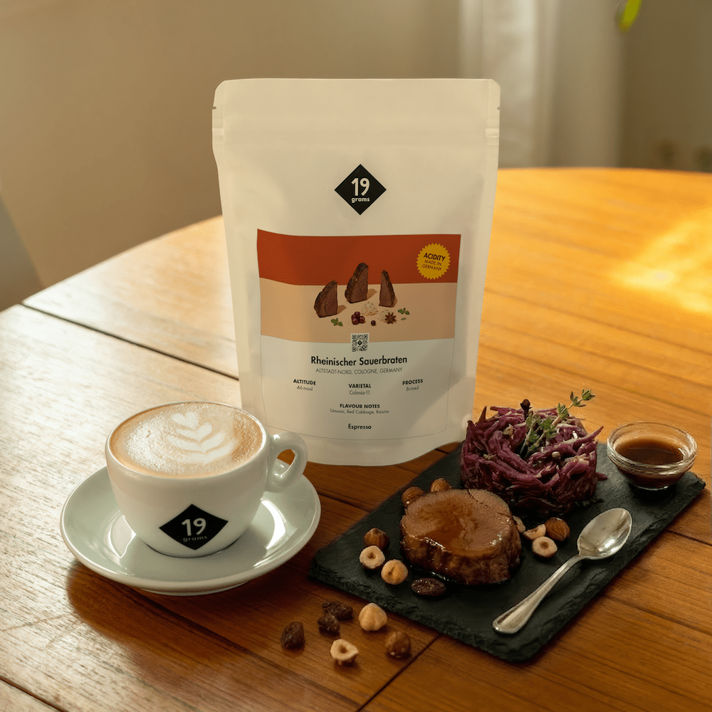 19grams Kaffeeverpackung „Rheinischer Sauerbraten" mit Cappuccino und Fleisch auf Holztisch