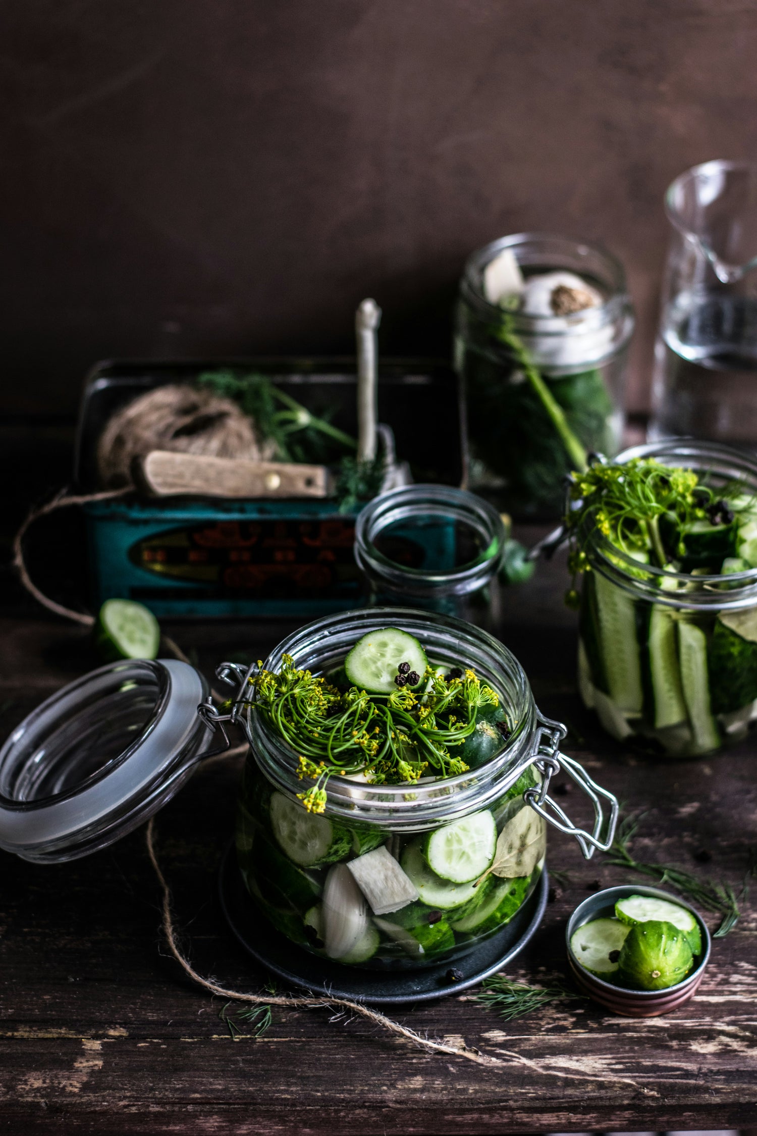 Einmachgläser mit eingelegten Gurkenscheiben, Dill und Kräutern auf dunklem Holztisch